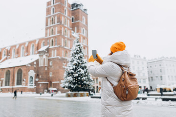 Attractive young female tourist is exploring new city. Redhead 30s woman in yellow hat resting on Market Square in Krakow. Traveling Europe in winter. St. Marys Basilica
