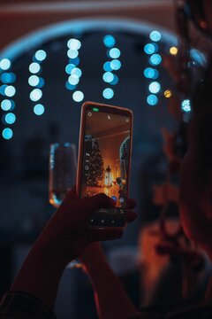 Person taking a festive photograph with their smartphone, surrounded by colorful Christmas lights