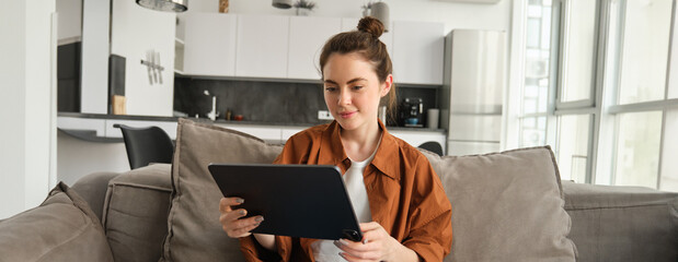 Portrait of young woman sitting on couch in living room, watching movies on digital tablet, reading on device, enjoying weekend at home