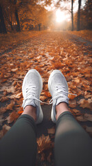 Feet of woman wearing white sneakers,  sitting on the ground in an autumn park. Beautiful autumn road view on background