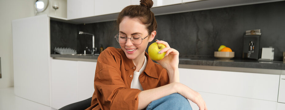 Beautiful young woman in glasses, sitting on chair in the kitchen, eating green apple and smiling