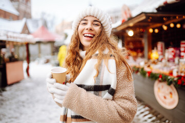 Happy woman drinks a hot drink from a glass and walks through the Christmas market on a sunny...
