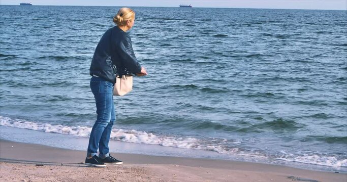 Blonde girl in jeans on the shore of the Black Sea throws pebbles into the sea against the backdrop of ships in the roadstead