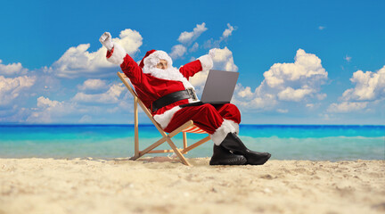 Santa claus at the beach sitting on a deck chair with a laptop computer