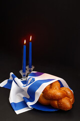 Challah bread covered with the Israeli flag, burning blue candles against a black background. Traditional Jewish Shabbat ritual. Shabbat Shalom.