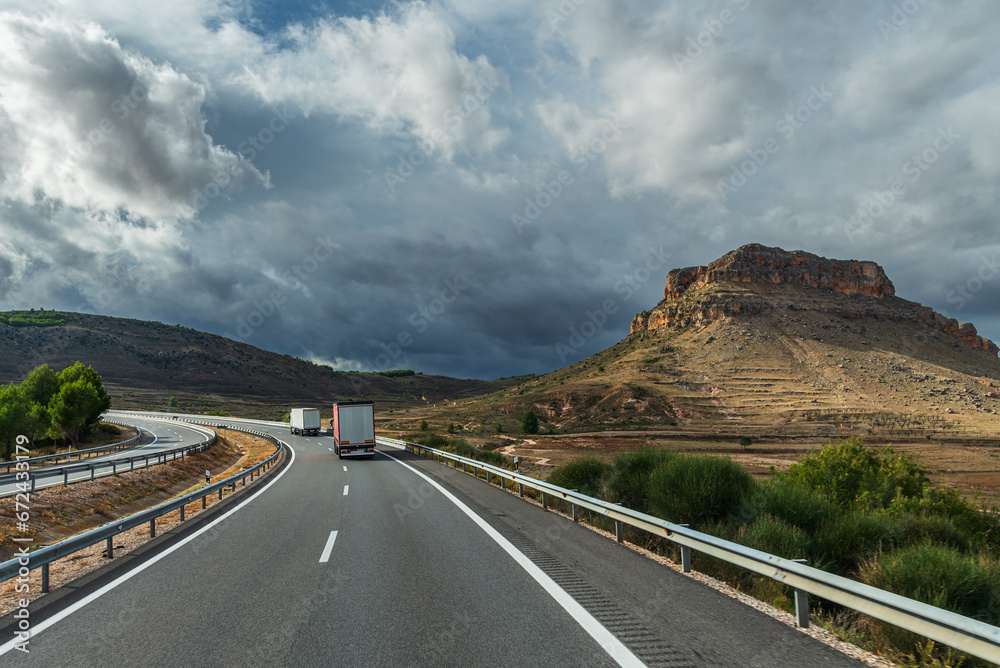 Wall mural trucks driving on a highway next to a rocky mountain and a sky that threatens a storm