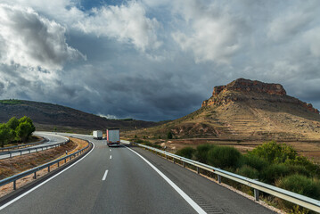 Trucks driving on a highway next to a rocky mountain and a sky that threatens a storm