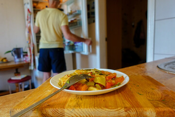 A plate of food on the table. In the background, a man opened the refrigerator.
