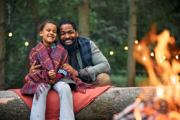 Happy black father and daughter enjoy by bonfire while camping in woods.