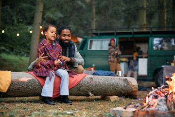 Black little girl and her father relax by campfire at trailer park.