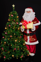 Santa Claus stands next to the Christmas tree and offers a boxes with a gifts. On the black background.