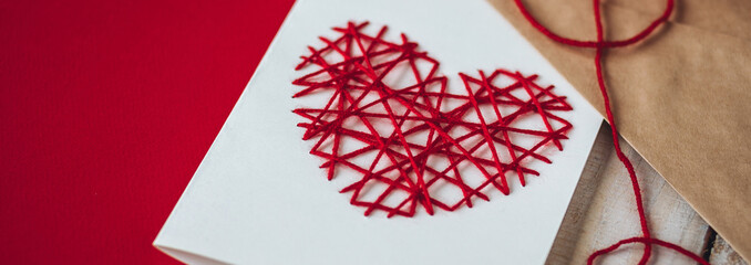 Concept of handmade simple holiday surprised for Saint Valentine's Day or Mother's Day. Easy to do DIY craft postcard from recycled paper with embroidering of red heart with wooden threads banner
