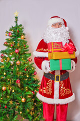 Santa Claus holds boxes with gifts next to a decorated Christmas tree.