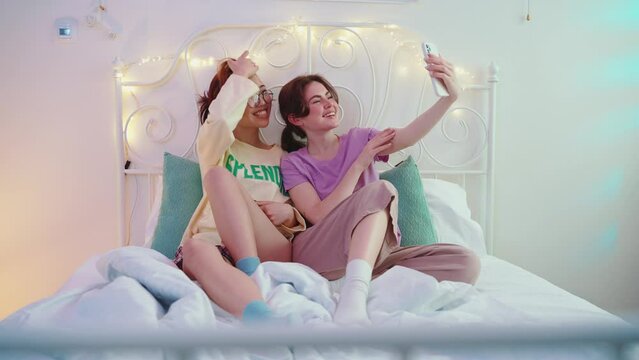 Two handsome multicultural female friends taking selfies on cellphone on the bed at a sleepover