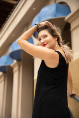 A beautiful girl in a black dress poses on the stairs against the background of beige buildings