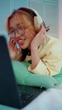 Funny young asian woman in eyeglasses watching video clip on laptop with headphones and singing on the bed at home