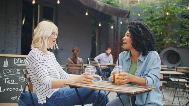 Positive multi-ethnic friends drinking cocktails in street cafe. Two girls speaking with each other while resting together in terrace. Gossiping or actively discussing something. Concept of meeting.