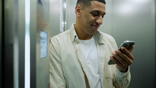 Positive african man texting on phone while riding in office elevator