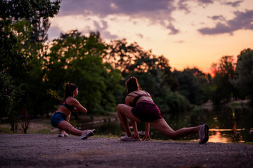 Fit girls working out at night in a green park, stretching and warming up their athletic bodies. Nature and a small lake provide the backdrop for their sporty training routine.