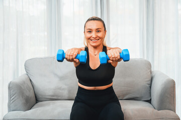 Fototapeta na wymiar Athletic and sporty senior woman engaging in body workout routine, sitting on sofa and lifting dumbbell at home as concept of healthy fit body with body weight lifestyle after retirement. Clout