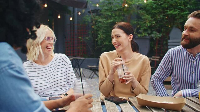 Friendly mixed-raced people having party at cafe on cozy terrace. Group of young women and men having funny conversation while drinking juice in summer day. Loudly laughing and joking together.
