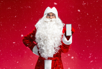 Man wearing Santa Claus costume  holding mobile phone showing device  screen looking at camera isolated on red background, mockup. Online shopping, sale, Christmas concept