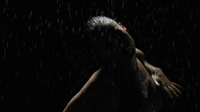 Silhouette elegant female dancing posing under rain at night. Seductive woman, gymnast dancer wearing black swimsuit, raindrops splashes, water drops roll down sexy body in shower, black background