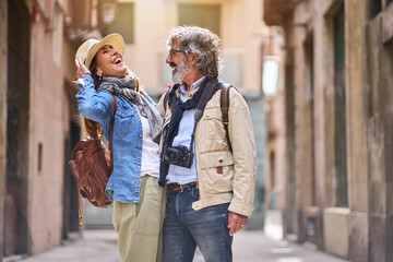 Laughing adult Caucasian couple standing embraced affectionately in tourist street of town. Mature people in love enjoying romantic vacation time. Positive relationships and weekend getaways spring. - obrazy, fototapety, plakaty