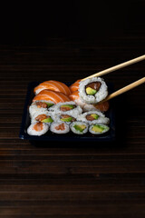 Assorted Takeout Sushi Tray. Holding Salmon Roll with Chopsticks . Wood Background.