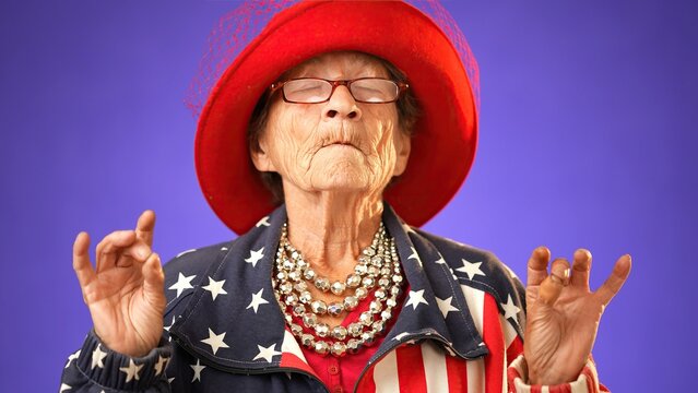 Funny toothless grandmother senior woman saying OM, 80s, meditating doing yoga exercise wearing US flag jacket isolated on purple background. Concept of youthful old female.