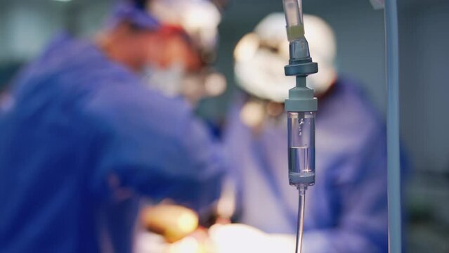 Drop counter reservoir half-filled with saline solution. Surgeons operate at backdrop in blur.