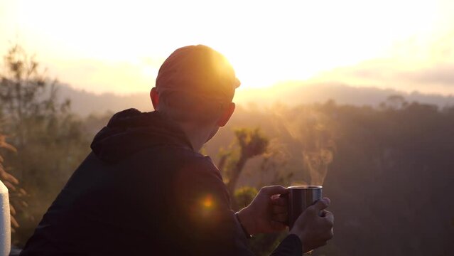 Hiker sitting on cliff near camping van against background of sunrise, holding hot mug with steam Close-up of man in hoodie in cold early morning watching sunrise Travel on camping van to meet sunrise