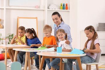 Nursery teacher and group of cute little children making toys from color paper at desks in kindergarten. Playtime activities