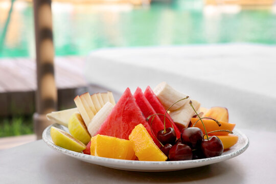 Plate with fresh fruits on table near sun lounger. Luxury resort with outdoor swimming pool