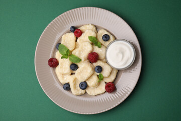 Plate of tasty lazy dumplings with berries, mint leaves and sour cream on dark green background, top view