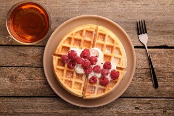 Tasty Belgian waffle with fresh raspberries, whipped cream, cup of tea and fork on wooden table, flat lay