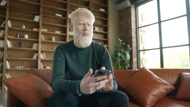 Focused man with blond hair frowning brows while sitting on leather sofa using phone. Serious concentrated Albino guy scrolling news, reading emails in modern urban loft apartment on motion background