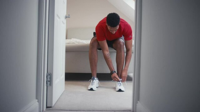 View through bedroom door of man in fitness clothing tying laces on trainers and checking fitness app on smart watch before exercise - shot in slow motion