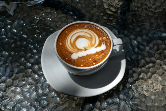 A cup of coffee with milk stands on a glass table in the sun in a street cafe. A snail is painted on the milk foam.