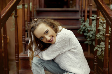 Fototapeta premium Young Gen Z woman sitting on the stairs in a spacious living room in a country house, looking at the camera and smiling. The concept of time outside the city, inner joy, calmness and good moments