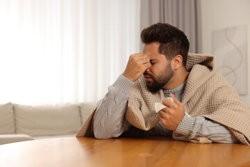 Sick man wrapped in blanket with tissue at wooden table indoors, space for text. Cold symptoms
