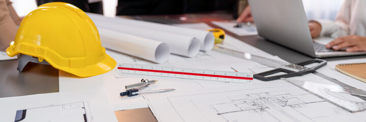 Engineer tool, architecture blueprint design and safety helmet hardhat on meeting table with contractors paper project with blurred engineer and architect working and discussing in background. Insight