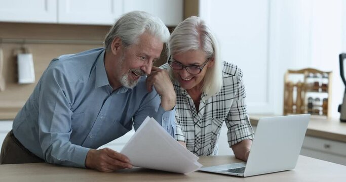 Cheerful positive older married couple reviewing document at laptop, discussing financial report from bank, reading paper insurance agreement, getting good news, investment income