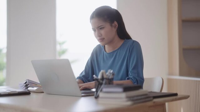 An Asian woman in loungewear looks concerned about her job and seeks solutions for a project she's struggling with. She uses her laptop at the dining table.