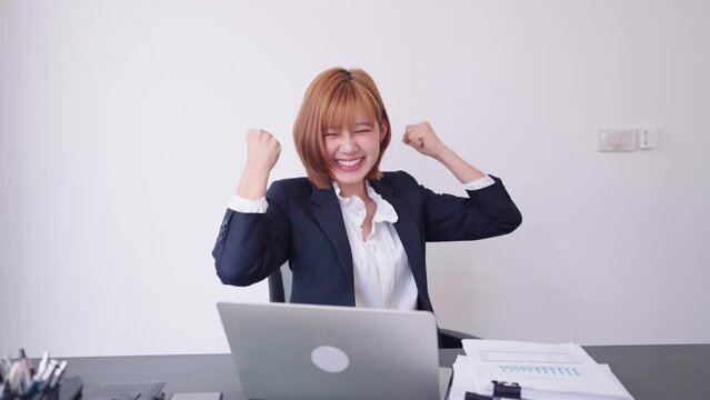 A cheerful Asian businesswoman, brimming with confidence, celebrates as her work receives support and recognition from her company at her excitedly prepared workspace