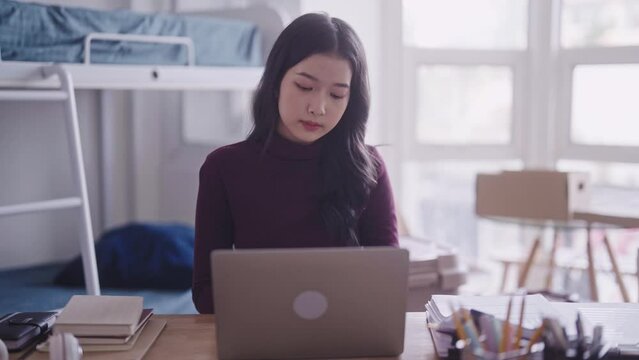 A diligent Asian female student, in casual attire, can be seen completing her homework or typing a research project or thesis on her laptop at her dormitory desk.