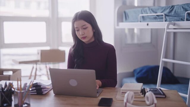 A diligent Asian female student, in casual attire, can be seen completing her homework or typing a research project or thesis on her laptop at her dormitory desk.