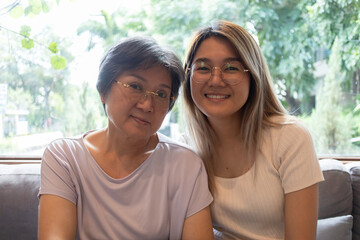Obraz premium Asian Thai old mother and daughter wear glasses, both woman happy smiling and looking at camera, sitting on sofa together, happy life, good family relationship concept.