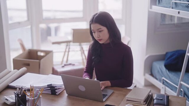 A diligent Asian female student, in casual attire, can be seen completing her homework or typing a research project or thesis on her laptop at her dormitory desk.