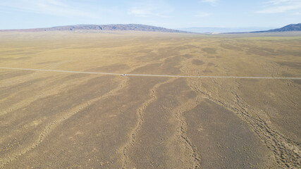 A white SUV is driving fast on a dusty road in the steppe. Clouds of dust fly from under the wheels and from the roof of the car. The jeep easily passes a dirt road. Blue sky and white clouds.